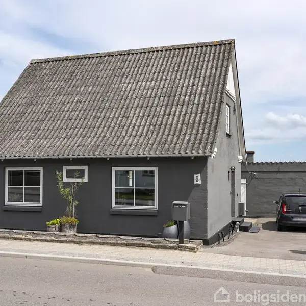 Gråt hus med skråtag står ved vejen, nummer fem på facaden. Ved siden af huset er en sort bil parkeret på indkørslen. Baggrund med grøn hæk og himmel.