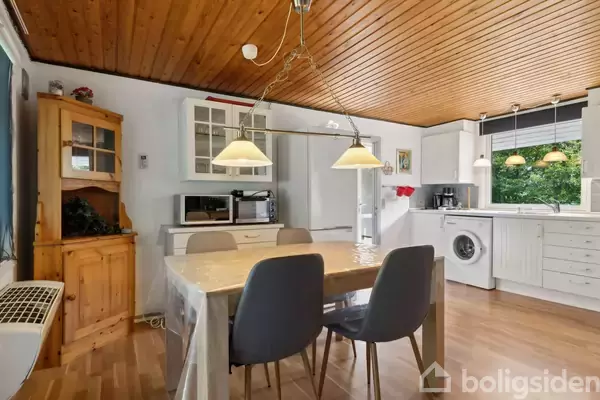 En spisebord dækket med plastikdug står i centrum, omgivet af grå stole. Over bordet hænger to lamper. I baggrunden er der køkkenelementer og en vaskemaskine i et lyst køkken.