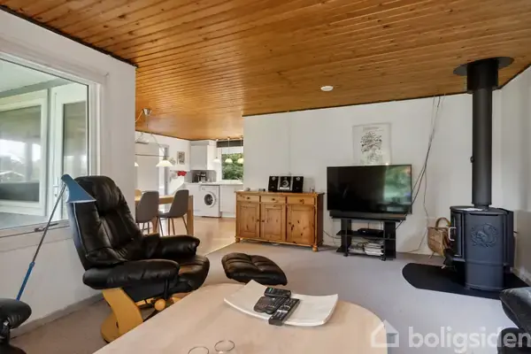 Stue med lænestole, fladskærms-tv på kommode, og brændeovn; køkken og spisestue ses i baggrunden. Træloft og hvide vægge skaber en hyggelig atmosfære.