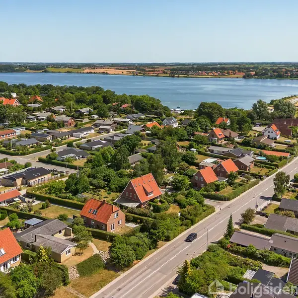 Huse med røde tage står stille i en frodig forstad ved en vej, nær en sø i baggrunden.