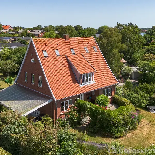 En stor murstensvilla med rødt tegltag står omgivet af en grøn, frodig have i et villakvarter. Der er flere vinduer og en overdækket terrasse.