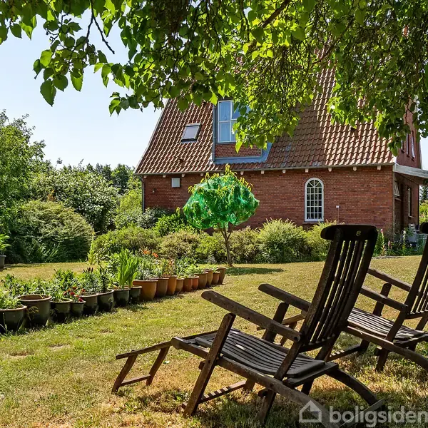 To træstole står på en græsplæne foran et rødt murstenshus med skråt tag. Huset er omgivet af frodig vegetation og flere krukker med planter.