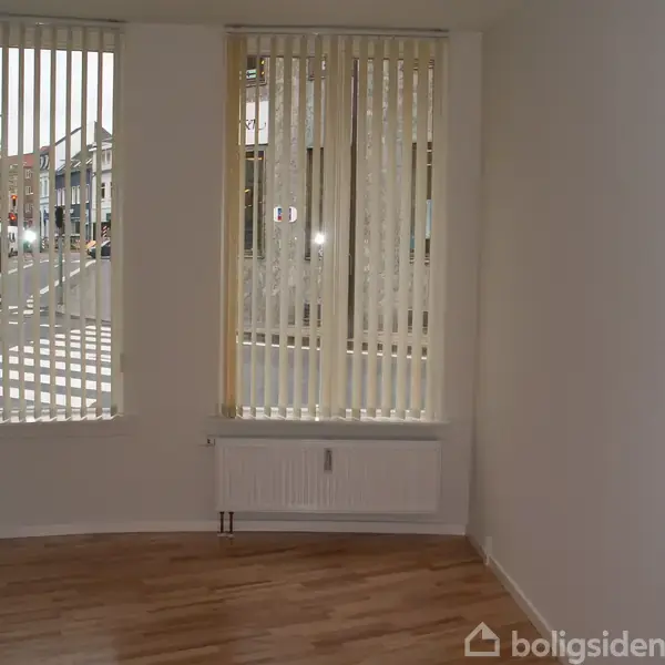 Et tomt rum med trægulv har store vinduer med vertikale persienner, der lukker lys ind. En radiator er placeret under vinduerne. Udenfor ses en gade med bygninger.