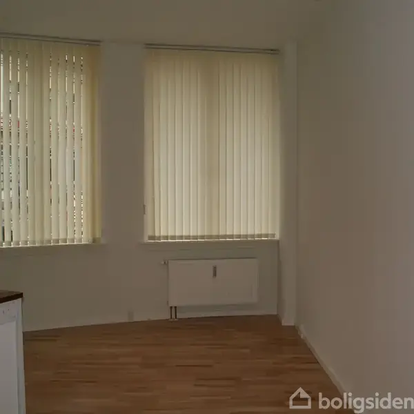En tom stue med lyse trægulve. To store vinduer med lodrette persienner dækker væggen, og der er en radiator under vinduerne. En del af et køkkenhjørne ses til venstre.