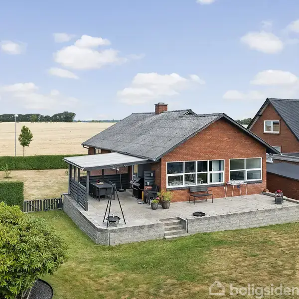 Huset står stille i en landlig have med en stor terrasse, omgivet af græsplæne og marker under en blå himmel.