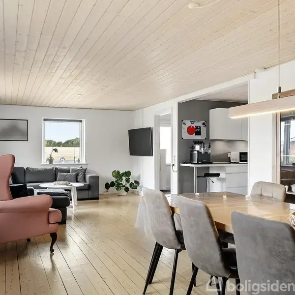 Lyst, moderne stue med trægulv og træloft. En rosa lænestol og en grå sofa står nær et vindue. Et spisebord med grå stole fylder rummet. Køkkenet ses i baggrunden.