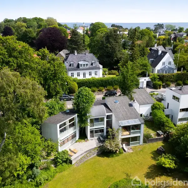 Flere moderne villaer ligger omgivet af frodige haver og store træer i et naturskønt boligkvarter. Husene har store vinduer og fladt tag, og området strækker sig mod en kystlinje.