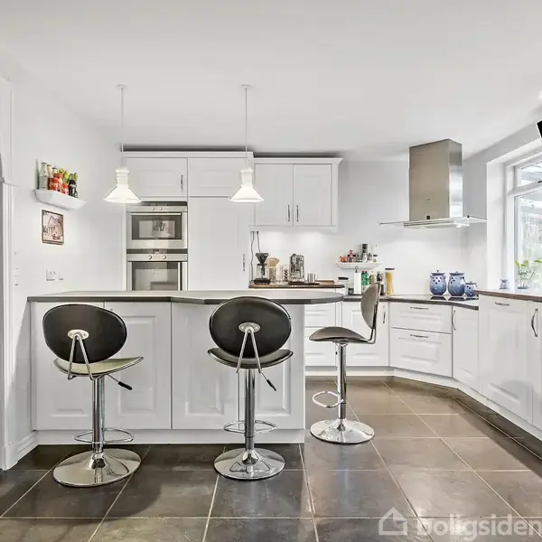 Et moderne køkken med hvidt interiør, tre barstole ved en central ø, omgivet af skabe og apparater. Der ses en åben buegang til en tilstødende stue med tæppe.