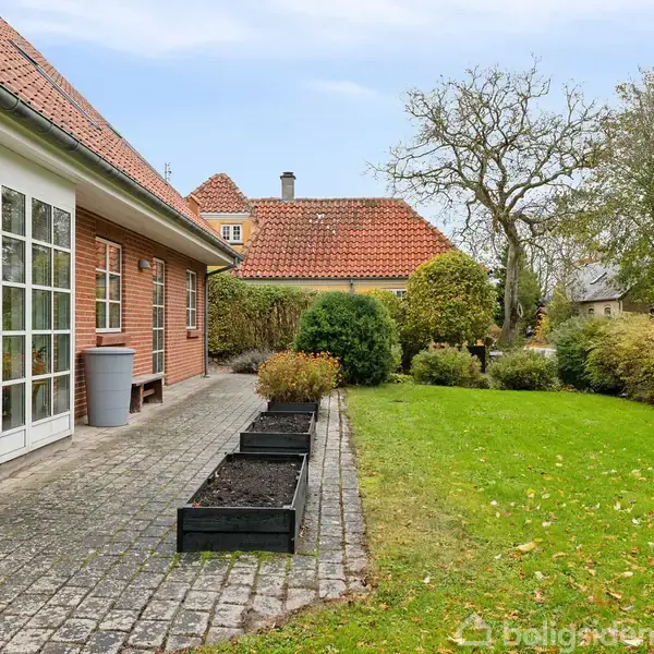 Rødt murstenshus med store vinduer på terrassen, flankeret af plantekasser. Ved siden af er der en velplejet have med grøn græsplæne og buske, omgivet af et større træ.