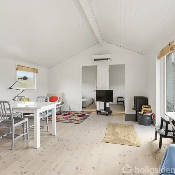 Stue med hvidt bord og stole i forgrunden, tv på væggen, og stor vindue med udsigt til have. Lysindfald skaber en rummelig atmosfære. Tæppet tilføjer farve.