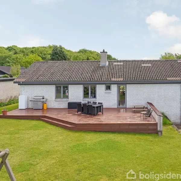 Hus med fladt tag og skorsten står i en grøn have. En træterrasse foran huset har spisebord og stole. En legeplads ses til venstre.