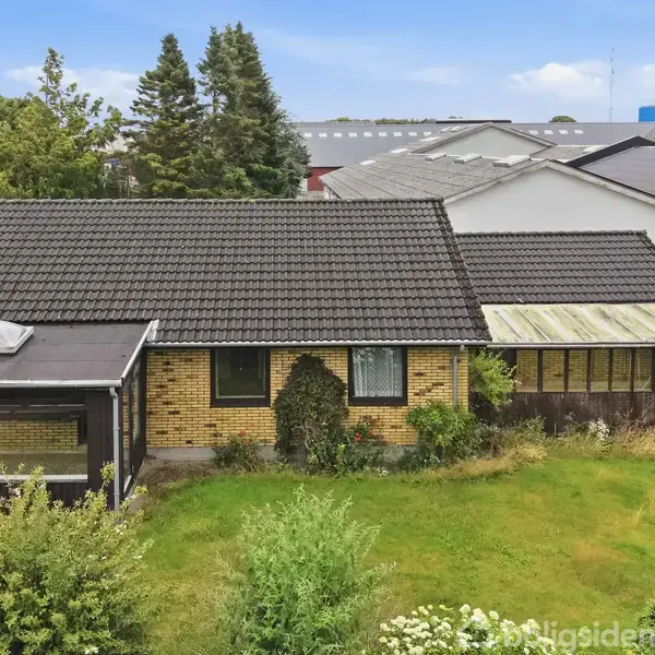 En gul murstensvilla med sort tag står stille omgivet af en grøn have. Det er beliggende i et villakvarter med andre huse i baggrunden.