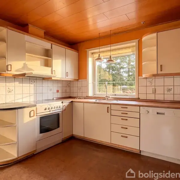Køkken med lyse skabe og hvidevarer; komfur og opvaskemaskine er integreret. Vinduet over vasken slipper naturligt lys ind. Omgivet af fliser og træloft. Ingen tekst til stede.