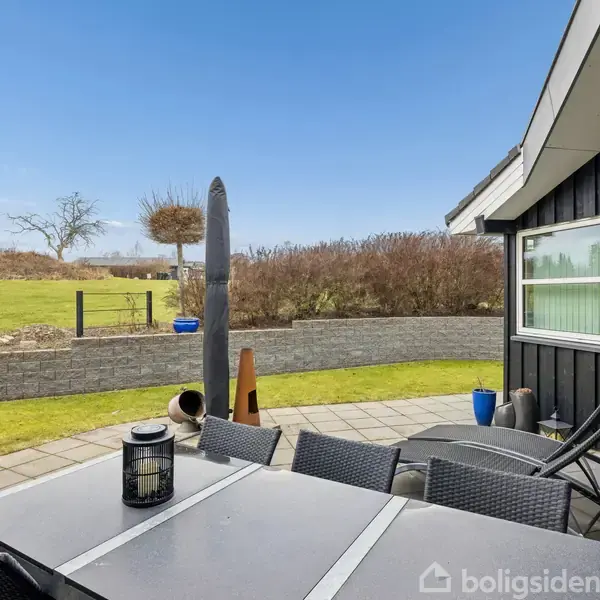 Terrassebord med stole står på flisebelagt terrasse ved siden af et sort træhus. I baggrunden er en grøn græsplæne og en lav mur, der adskiller terrassen fra haven.