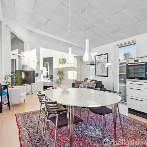 Ovalt spisebord i hvidt med sorte stole omkring, placeret på et blomstret tæppe i et lyst, moderne opholdsrum med højt til loftet. Mørk sofa og fladskærms-tv i baggrunden.