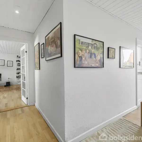 Gang indeholder vægge med indrammede billeder, der leder til et rum med store glasdøre. Træparketgulv breder sig gennem gangen, og gennem et vindue ses en have med græs og huse.