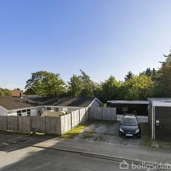 Bil parkeret i en indkørsel ved siden af et hus, omkranset af hegn. Træer fylder baggrunden under en klar blå himmel. Der ses en stille vej foran ejendommen.