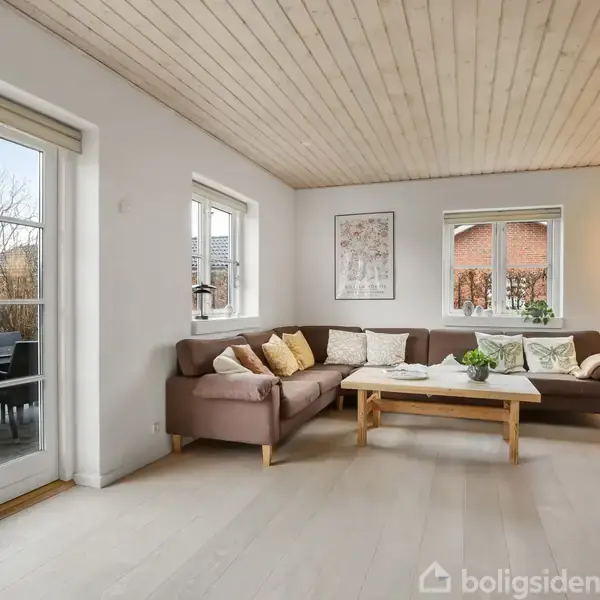 Brun sofa med puder står i en lys stue. Træbord foran sofaen. Store vinduer slipper lys ind. Dør til terrasse med udemøbler. Vægkunst og planter som dekoration.