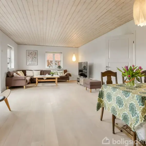 En rummelig stue med hvide vægge og lofter. Et spisebord med blomstret dug og vase står nær en hyggekrog med en sofa og lænestol. Store vinduer lader lyset strømme ind.