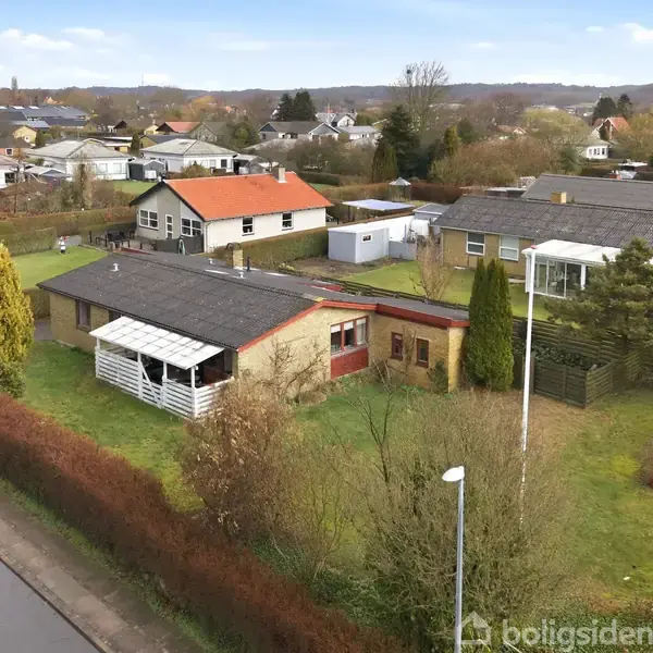En en-families hus med fladt tag og veranda står i en grøn have. Andre huse og træer omgiver det i et roligt boligkvarter.