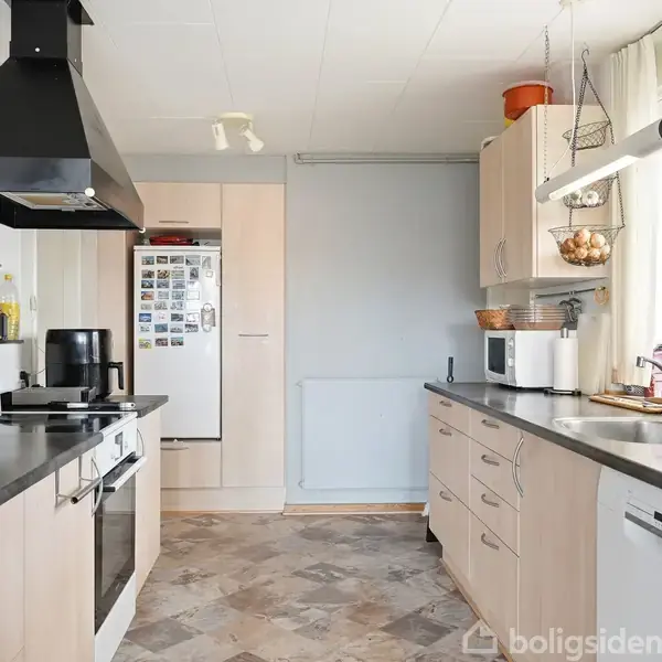 Køkken med lyse træskabe og grå bordplader. En kogeplade, emhætte og køleskab findes til venstre. Til højre står kaffemaskine og mikrobølgeovn. Vindue med gardiner bringer lys ind.