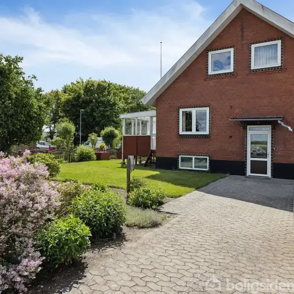 Et rødt murstenshus med store vinduer står i en velholdt have. En flisebelagt sti fører til indgangen. Flere buske og træer omkranser området under en blå himmel.