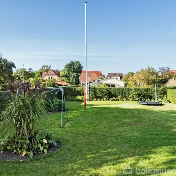 Flagstang står i en velplejet have med grøn græsplæne og buske. Haven er omgivet af hække, med flere huse i baggrunden under en klar blå himmel.