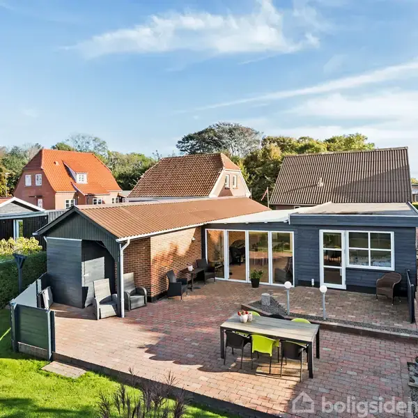 En moderne villa med rødt tag og stor glasfacade befinder sig i et villakvarter. Foran huset er en flisebelagt terrasse med et bord og stole.