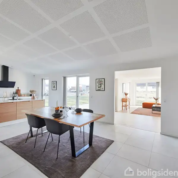 Et moderne køkken-alrum med et træbord og sorte stole på et tæppe. Rummets åbne design viser en stue med en sofa i baggrunden. Store vinduer bader rummet i lys.