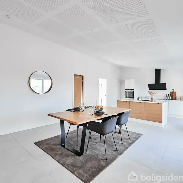 Et moderne køkkenalrum med et stort træbord i centrum, omgivet af sorte stole. I baggrunden ses et køkken med hvidt og træfarvet interiør, og rummet er lyst og rummeligt.