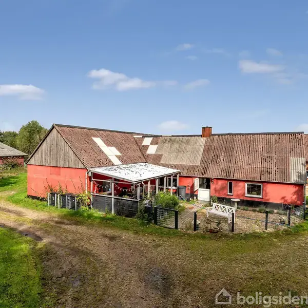 Rødt landhus med tag af falmede paneler og skyggefuldt verandaområde, placeret i landligt område med have og grusvej. Der er grønne marker og træer omkring huset.