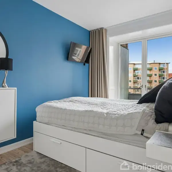 Soveværelse med hvid seng, placeret mod en blå væg. Væghængt skab med rund spejl, lampe, og fjernsyn. Store vinduer med udsigt til bygninger. Beige gardiner dækker delvist vinduerne.