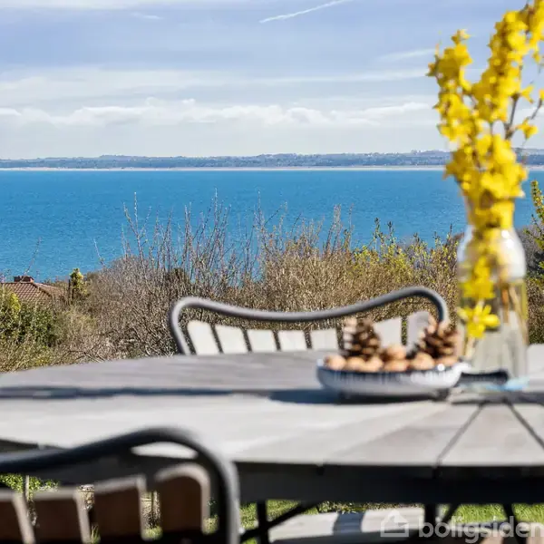 Et rundt bord står på en terrasse med en vase med gule blomster og en skål. I baggrunden ses en udsigt over buske og havet under en blå himmel.