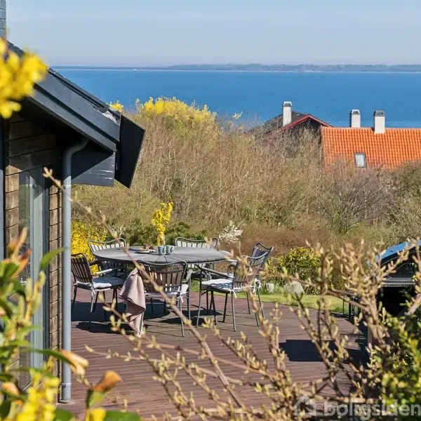 Hus med terrasse har havemøbler og udsigt over vand. Omgivet af buske og træer med rødt hustag i baggrunden. Klare blå himmel og hav skaber en rolig atmosfære.