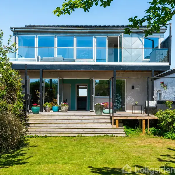Et moderne to-etagers hus med store glaspartier på øverste etage. En rummelig terrasse med potteplanter foran, omgivet af grønne træer og en velplejet plæne.