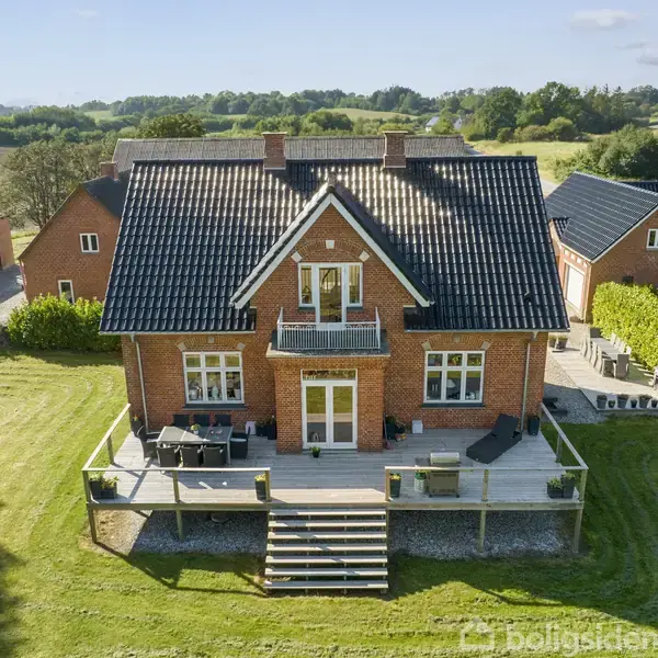 Rødt murstenshus med sort tag har en stor træterrasse med møbler og trapper. Huset ligger i en frodig, landlig omgivelser med marker og træer i baggrunden.