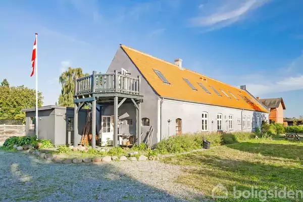 En grå bygning med orange tag står i en gård, omgivet af græsplæne og træer. En flagstang med Dannebrog er placeret til venstre, og en balkon er synlig på bygningens forside.