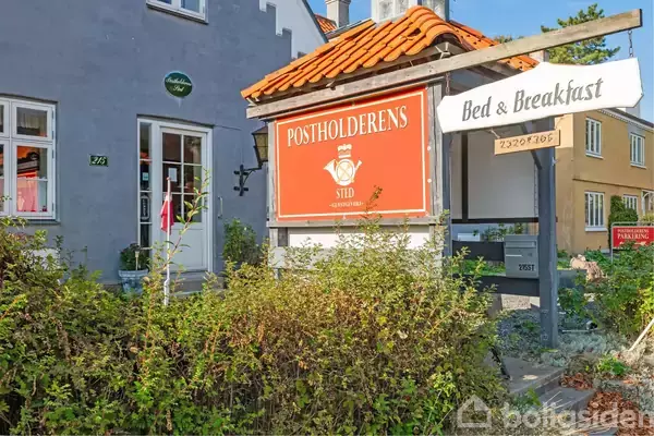 Et skilt med teksten "POSTHOLDERENS STED" hænger fra en træstruktur, mens det er placeret foran en bygning med en dør og vinduer. Omgivet af buske og grønne planter.