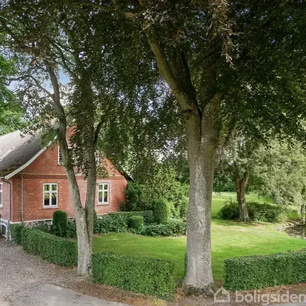 Et rødt murstenshus står stille omgivet af en veltrimmet have med tætte hække og træer. En grussti leder op til huset, mens en dam kan ses i baggrunden.