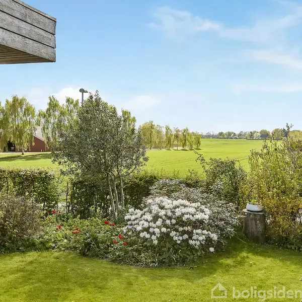 Haven med blomsterbede og buske, omgivet af en velplejet græsplæne, skaber en frodig udsigt mod fjerne træer og åbne marker under en blå himmel.