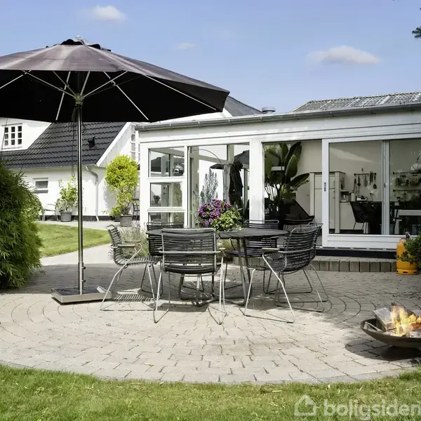 Terrasse med rundt bord og stole under en stor parasol. I baggrunden et hvidt hus med store vinduer. To ildsteder flankerer terrassen omgivet af grønne planter.