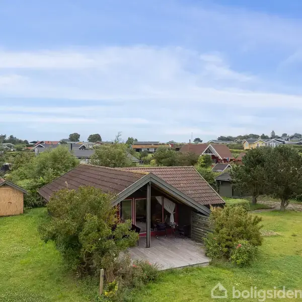 Et træhus med skråt tag står stille på en grøn græsplæne omgivet af buske og træer, beliggende i et område med andre huse i det fjerne.