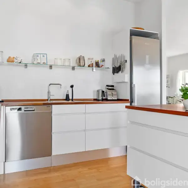 Et moderne køkken med hvide skabe og træbordplader. En opvaskemaskine indlejret i indretningen. På væggen er hylder med køkkenartikler, og i baggrunden ses en stue med en plante.