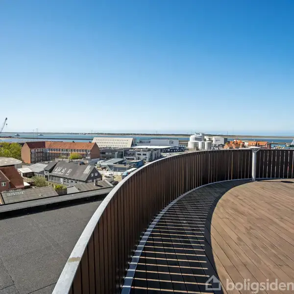 En rund tagterrasse med trædæk står stille, omkranset af en metalrækværk. I baggrunden ses bybygninger, industri og havn under en klar blå himmel.