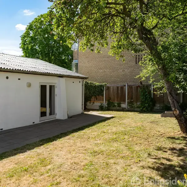 Et hvidt hus med store vinduer står til venstre, mens en frodig have med en stor træ dominerer højre side. Græsset er let tørt, og der er en stensat terrasse.