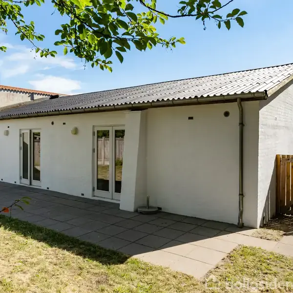 Et hvidt, enetages hus står stille i en solrig have. Husets overflade har store vinduer og en flisebelagt terrasse. Omkring huset er der græsplæner og træer.