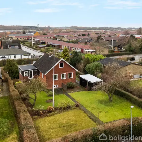 Rødt murstenshus med sort tag står stille i en velplejet have med buske og træer. Omkringliggende rækkehuse og en blå himmel former kvarteret.