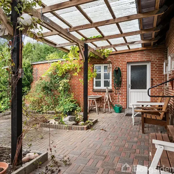 Terrasse med møbler under et overdækket tag. Vægge af rød mursten omgiver området. Krukker med planter står langs kanten, mens døren fører til et tilstødende rum.