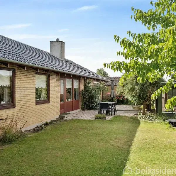En gul murstensvilla står i en velplejet have, hvor der er en terrasse med havemøbler. Omkring huset er der grønne træer og buske under en klar, blå himmel.