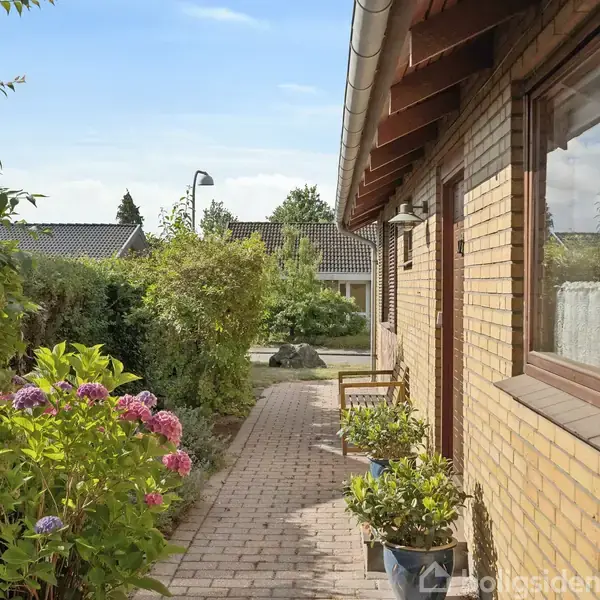 Murstenshus med havegange og blomsterkrukker, der fører til hoveddøren. Omgivet af buske og blomster, giver en hyggelig atmosfære i et villakvarter.